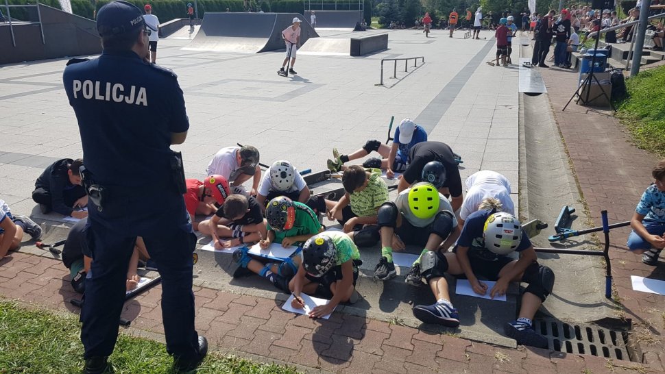 Skatepark w Bieruniu. Na pierwszym planie policjant stoi tyłem. Na drudim planie dzieci siedzą na boisku i rozwiązują quiz, obok leżą hulajnogi. W tle osoby korzystające z parku.