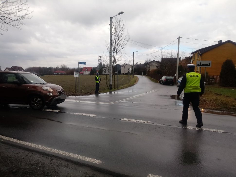 Policjanci zatrzymują do kontroli drogowej samochód osobowy w kolorze brązowym. Jeden policjant w kamizelce odblaskowej stoi na jezdni tyłem (po prawej stronie). Na drugim planie stoi drugi policjant. W tle widać zabudowania.