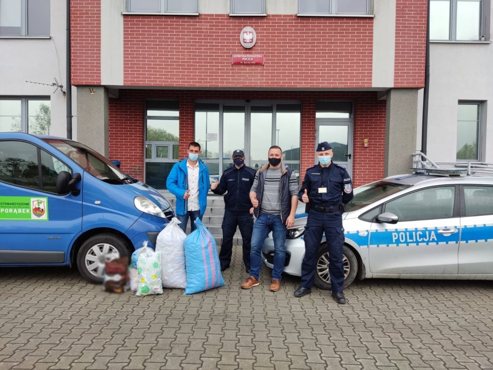 Przed wejściem do komendy w Bieruniu stoją dwaj policjanci i dwaj przedstawiciele stowarzyszenia pomiędzy zaparkowanymi pojazdami - radiowozem policyjnym i samochodem w kolorze błękitnym. Na pierwszym planie stoją worki z nakrętkami.