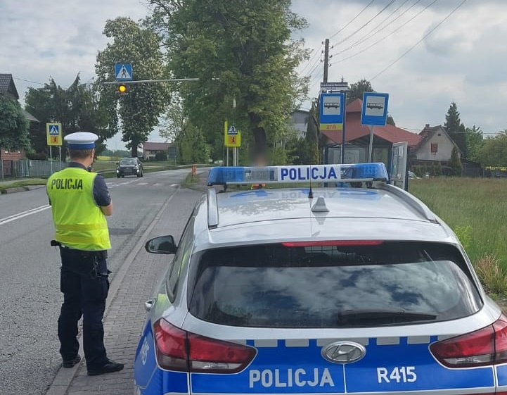 Policjant ruchu drogowgo w odblaskowej kamizelce stoi przy radiowozie na poboczu ulicy, w rejonie przejścia dla pieszych