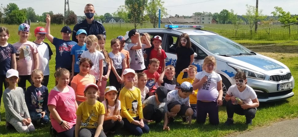 Grupa dzieci uczestniczących w spotkaniu i policjant stoją przy radiowozie. Część dzieci klęczy na trawie. W tle widać drzewa i zabudowania