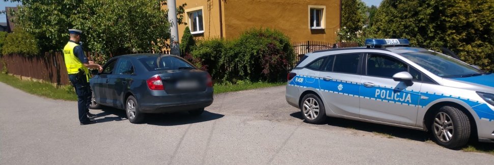 W lewej części zdjęcia policjant ruchu drogowego stoi przy zatrzymanym do kontroli samochodzie w kolorze granatowym, w pawej częśći stoi zaparkowany radiowóz. W tle widać fragment budynku