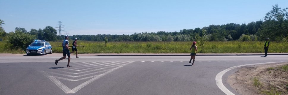 Trasa biegu. Uczestnicy biegną ulicą, na poboczu stoi radiowóz. W prawej części fotografii policjant ruchu drogowego stoi na jezdni i zabezpiecza bieg.