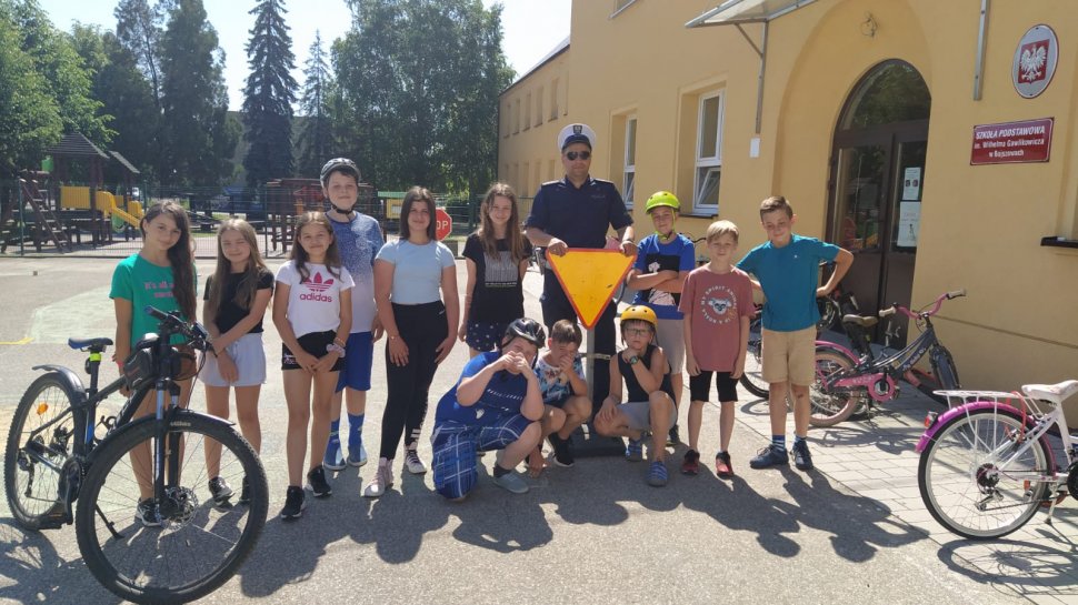 Przed szkołą w Bojszowach policjant stoi z grupą dzieci, które zdawały egzamin na kartę rowerową. Obok stoją rowery, policjant trzyma żółty znak drogowy. W tle widać budynek szkoły i drzewa.