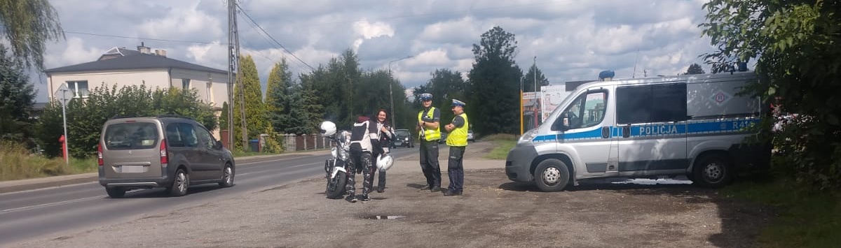 Policjanci ruchu drogowego w trakcie działań, stoją przy jezdni, obok stoi motocykl, jego kierowca i pasażer. Po prawej stronie widać zaparkowany radiowóz. Jezdnią przejeżdża samochód. W tle widać zabudowania i drzewa.