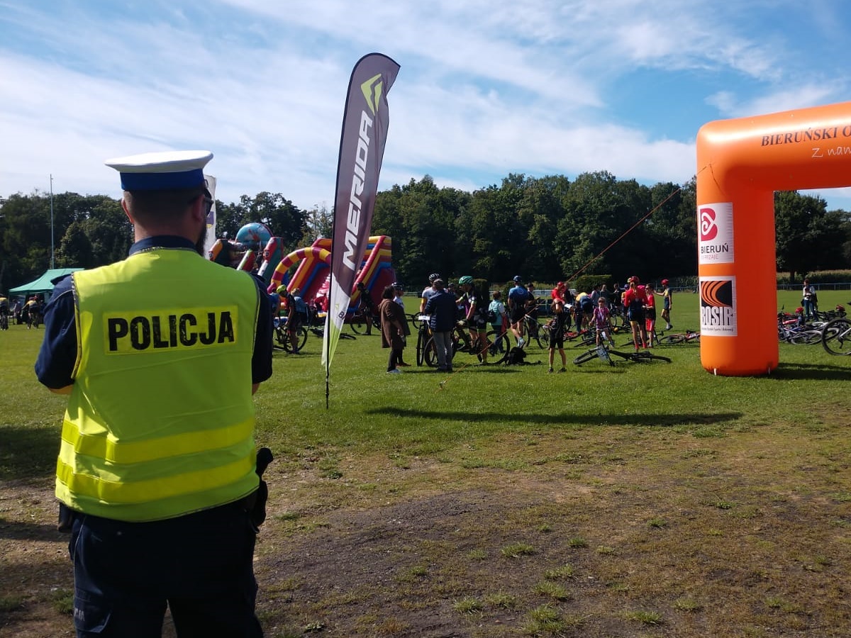 Na pierwszym planie policjant ruchu drogowego w odblaskowej kamizelce stoi tyłem. Na drugim planie widać uczestników rajdu, którzy stoją z rowerami w rejonie miejsca wyznaczonego na start