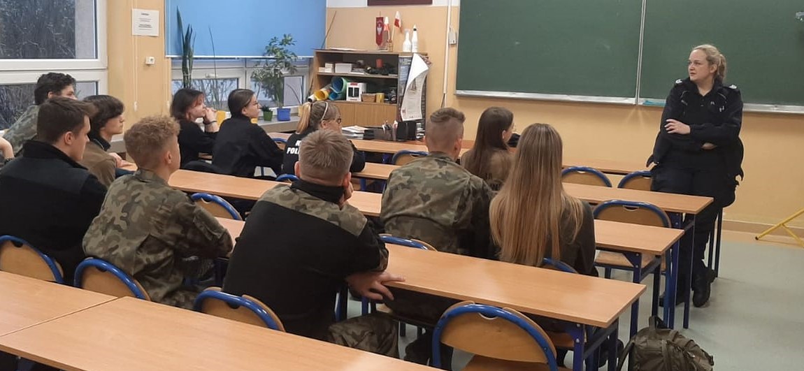 W sali lekcyjnej policjantka stoi pod tablicą, przodem do uczniów, którzy siedzą w ławkach. Policjantka prowadzi prelekcję. Uczniowie maja na sobie mundury.