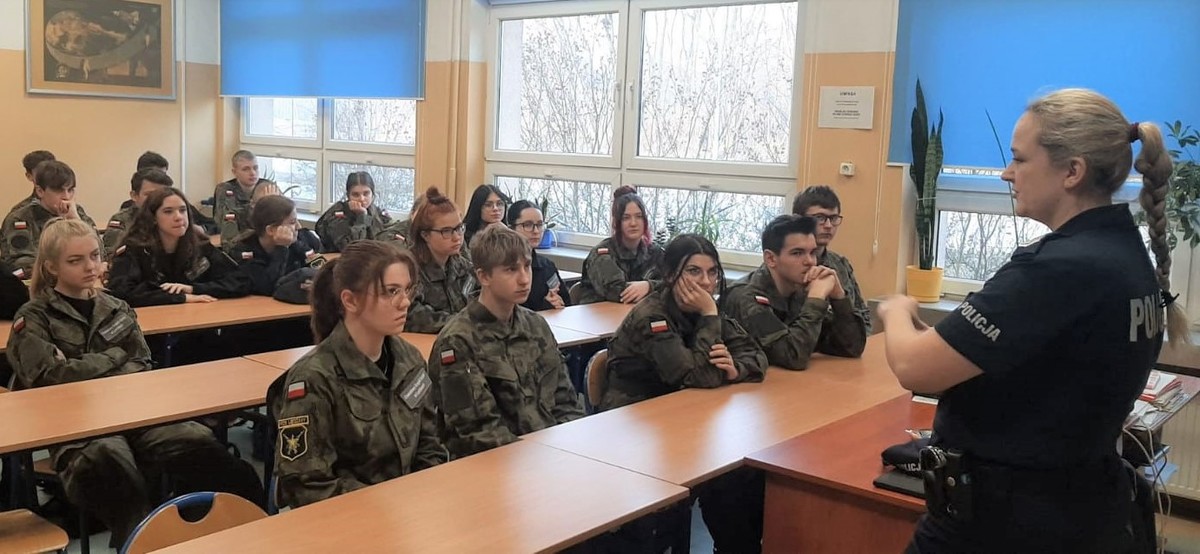 W sali lekcyjnej policjantka stoi przodem do uczniów, którzy siedzą w ławkach. Policjantka prowadzi prelekcję. Uczniowie maja na sobie mundury. Na drugim planie widać okna.