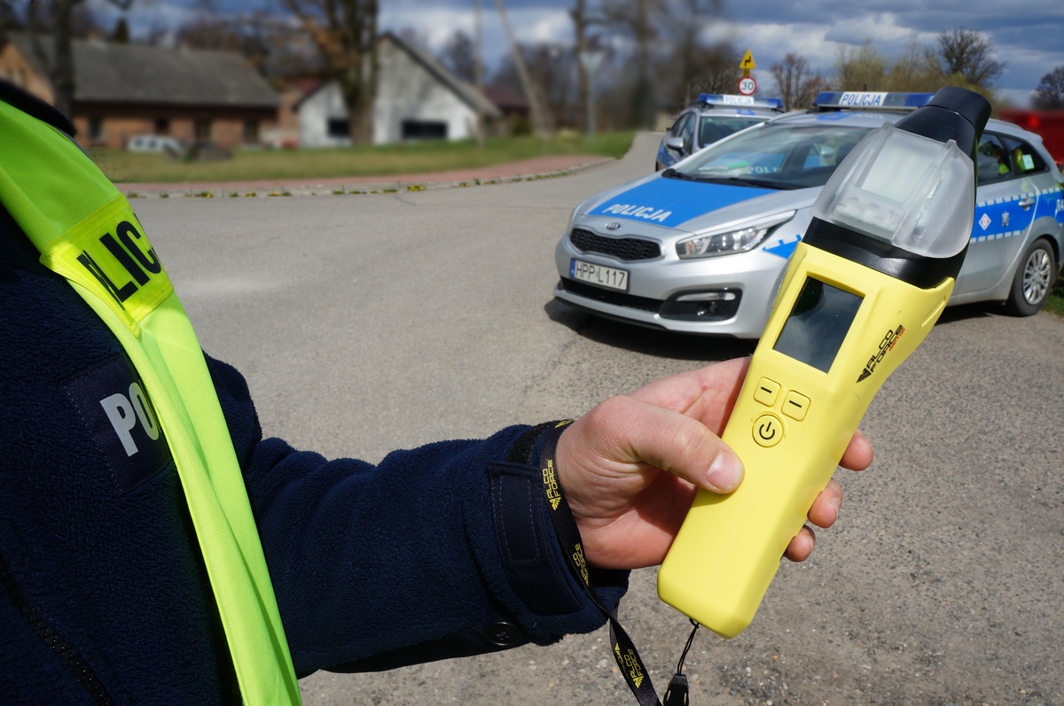 Na zdjęciu widzimy radiowóz policyjny oraz policjanta trzymającego urządzenie do badania stanu trzeźwości kierujących