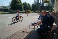 W lewej części zdjęcia dziecko na rowerze, pop prawej stronie policjanci i nauczycielka siedzą przy stoliku