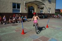 Na pierwszym planie dziewczynka na rowerze w trakcie egzaminu na kartę rowerową, na drugim planie dzieci siedzą na ławce, policjanci i nauczycielka siedzą przy stoliku, rowery stoją pod ścianą szkoły