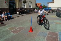 Na pierwszym planie chłopiec na rowerze, na drugim planie dzieci siedzą na ławce, policjanci i nauczycielka siedzą przy stoliku, rowery stoją pod ścianą szkoły