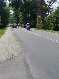 Drogą asfaltową na czele jedzie motocyklista, a za nim jedzie liczna grupa kolarzy.
