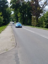 Po drodze jedzie oznakowany radiowóz policyjny zabezpieczający przejazd kolarzy, za nim jadą samochody z obsługi technicznej wyścigu kolarskiego.