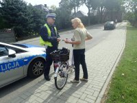 Kobieta z rowerem stoi przy policjancie, po lewej stronie widać część radiowozu, w tle samochód na ulicy