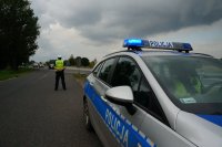 Na pierwszym planie w prawej części zdjęcia radiowóz policyjny. Na drugim planie przy jezdni stoi policjant ruchu drogowego.