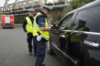 Dwaj policjanci ruchu drogowego stoją przy zatrzymanym do kontroli drogowej samochodzie. W tle widać część wozu strażackiego i most kolejowy.