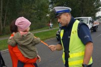 Policjant ruchu drogowego (po prawej stronie) wręcza opaskę odblaskową dziecku trzymanemu na rękach przez kobietę (po lewej stronie).