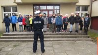 Na pierwszym planie stoi policjant tyłem, na drugim planie dzieci stoją przy wejściu do szkoły