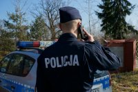 Policjant stoi przy radiowozie, trzyma telefon przy uchu