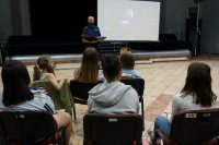 Na pierwszym planie dzieci siedzą na krzesłach, widać je od tyłu. Na drugim planie stoi policjant.