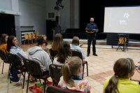 Na pierwszym planie dzieci siedzą na krzesłach, widać je od tyłu. Na drugim planie stoi policjant.