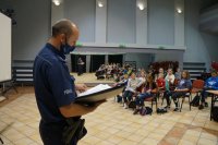 Na pierwszym planie stoi policjant w maseczce, trzyma notatnik. Na drugim planie dzieci siedzą na krzesłach.