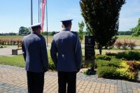Policjanci stoją przy obelisku, widać ich od tyłu. W tel flaga biało-czerwona na maszcie