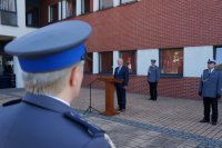 Na pierwszym planie, wlewej części fotografii widać fragment policjantki, na drugim planie stoją - starosta przy mównicy i policjanci