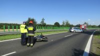 miejsce wypadku drogowego na pierwszym planie policjant wraz z strażakiem pożarnym wykonujący czynności przy motocyklu na drugim planie radiowóz oraz wóz strażacki