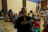 Na pierwszym planie policjantka stoi tyłem, na drugim planie dzieci siedzą na pufach
