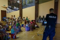 Na pierwszym planie w prawej części zdjęcia policjant stoi tyłem, na drugim planie dzieci siedzą na pufach