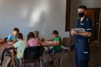 Po prawej stronie stoi policjant, po lewej dzieci siedzą przy stołach