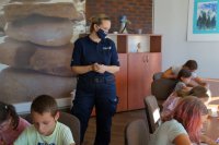 W centralnej części zdjęcia stoi policjantka, wokół dzieci siedzą przy stołach