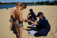 Plaża na Łysinie, w lewej części zdjęcia stoją dwaj chłopcy w strojach kąpielowych, w prawej części policjanci kucają policjantka wręcza dzieciom ulotki. W tle widać zalew