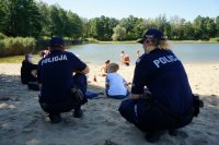 Plaża na Łysinie, na pierwszym planie policjanci kucają, na drugim planie dzieci siedzą na piasku, w tle widać plażowiczów i zalew