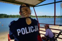 Punkt ratowników na plaży, na pierwszym planie policjantka stoi tyłem, na drugim planie ratownik - kobieta sidzi na fotelu plażowym, w tle widać zalew