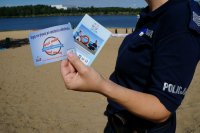 Policjantka trzyma ulotki kampanii &quot;Kręci mnie bezpieczeństwo nad wodą&quot; - widać tylko korpus osoby w prawej części zdjęcia. W tle plaża i zalew