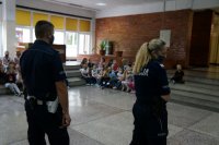 Na pierwszym planie stoją policjanci, na drugim planie dzieci siedzą na schodach szkolnej auli