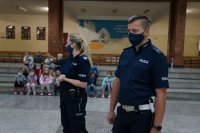 Na pierwszym planie stoją policjanci, na drugim planie dzieci siedzą na schodach szkolnej auli