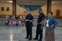 Na pierwszym planie stoją policjanci i pedagog szkolny, na drugim planie dzieci siedzą na schodach szkolnej auli