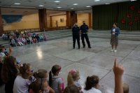Na szkolnej auli policjanci prowadzą prelekcję, obok stoi pedagog szkolny, dzieci siedzą na schodach