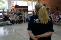 Na pierwszym planie policjantka stoi tyłem, na drugim planie dzieci siedzą na schodach szkolnej auli