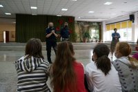 Na pierwszym planie widać od tyłu siedzące dzieci, na drugim planie stoją policjanci i szkolny pedagog