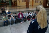 Po prawej stronie policjantka stojąca tyłem, na drugim planie dzieci siedzą na schodach szkolnej auli