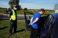 Działania Road Safety Days na DK44 w Bieruniu, mężczyzna w niebieskiej koszulce stoi przy samochodzie i słucha udzielanego mu przez strażaka instruktażu, po lewej stronie stoi policjant i przygląda się