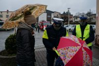 Policjanci wręczają odblaski kobiecie i dziecku, którzy trzymają kolorowe parasolki.