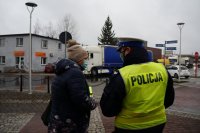 Policjant rozmawia z kobietą, która trzyma opaskę odblaskową. Oboje stoją przodem do ulicy, po której jadą samochody.