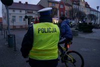 Policjant stojący tyłem rozmawia z rowerzystą. W tle widać policyjny radiowóz i wóz strażacki