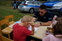 Policjant i chłopiec siedza przy stoliku na trawie. W tle stoją radiowozy